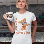 A woman standing in a kitchen holding a white mug with a colorful graphic and text, wearing a white graphic T-shirt featuring an illustrated muscular figure and the words 'OWAIN GLYNDWR'.