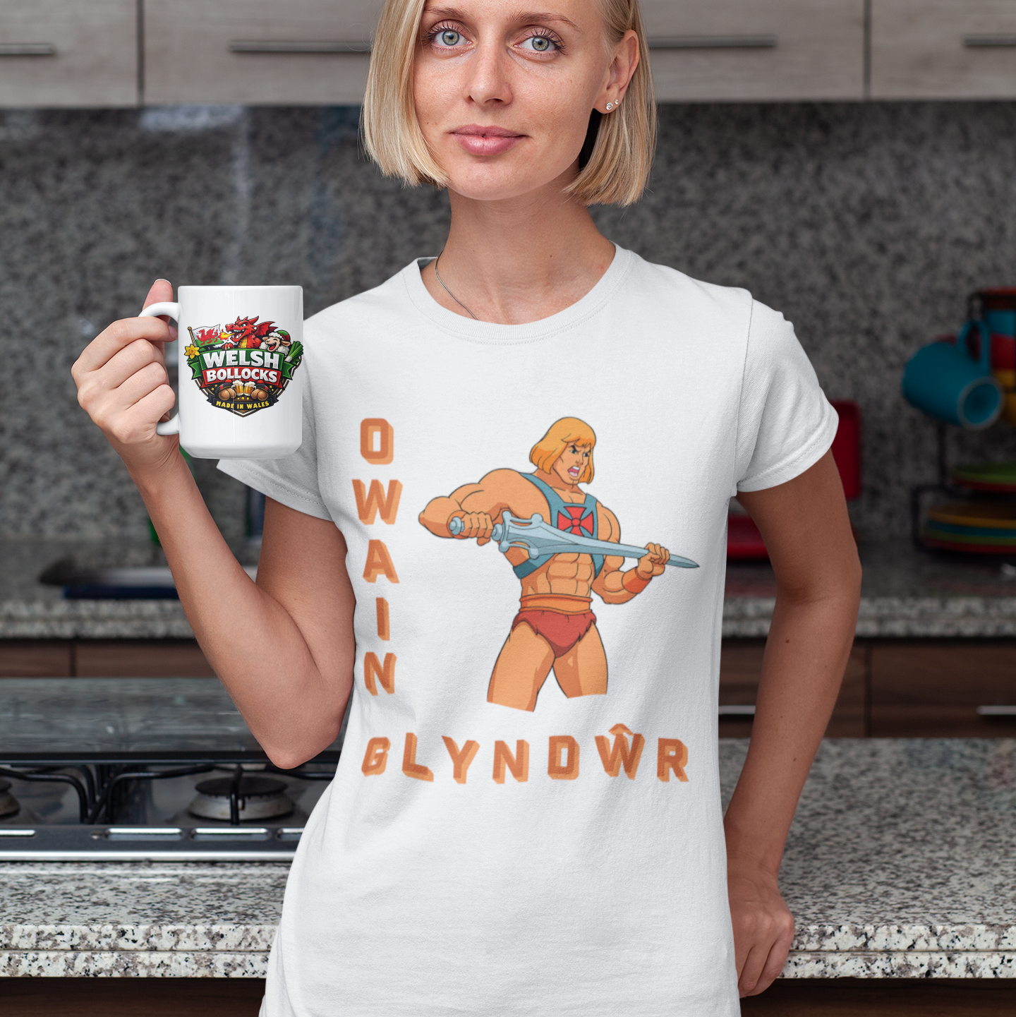 A woman standing in a kitchen holding a white mug with a colorful graphic and text, wearing a white graphic T-shirt featuring an illustrated muscular figure and the words 'OWAIN GLYNDWR'.