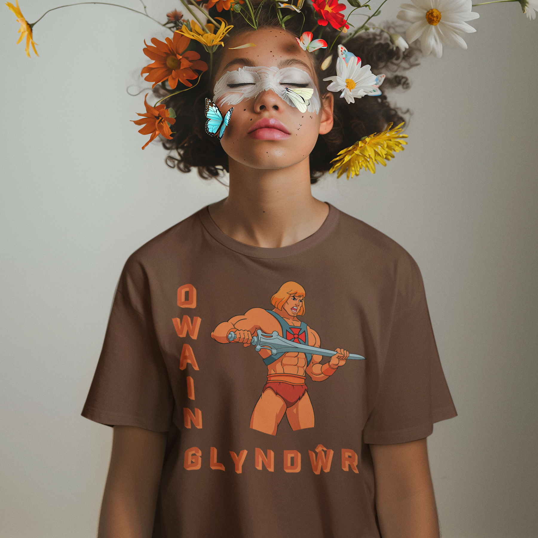 A person stands against a plain light gray background wearing a brown graphic T-shirt with a colorful flower arrangement and butterflies floating around the head area. The face is obscured by a large rectangular blur/block, and the shirt graphic includes stylized text and an illustrated figure.