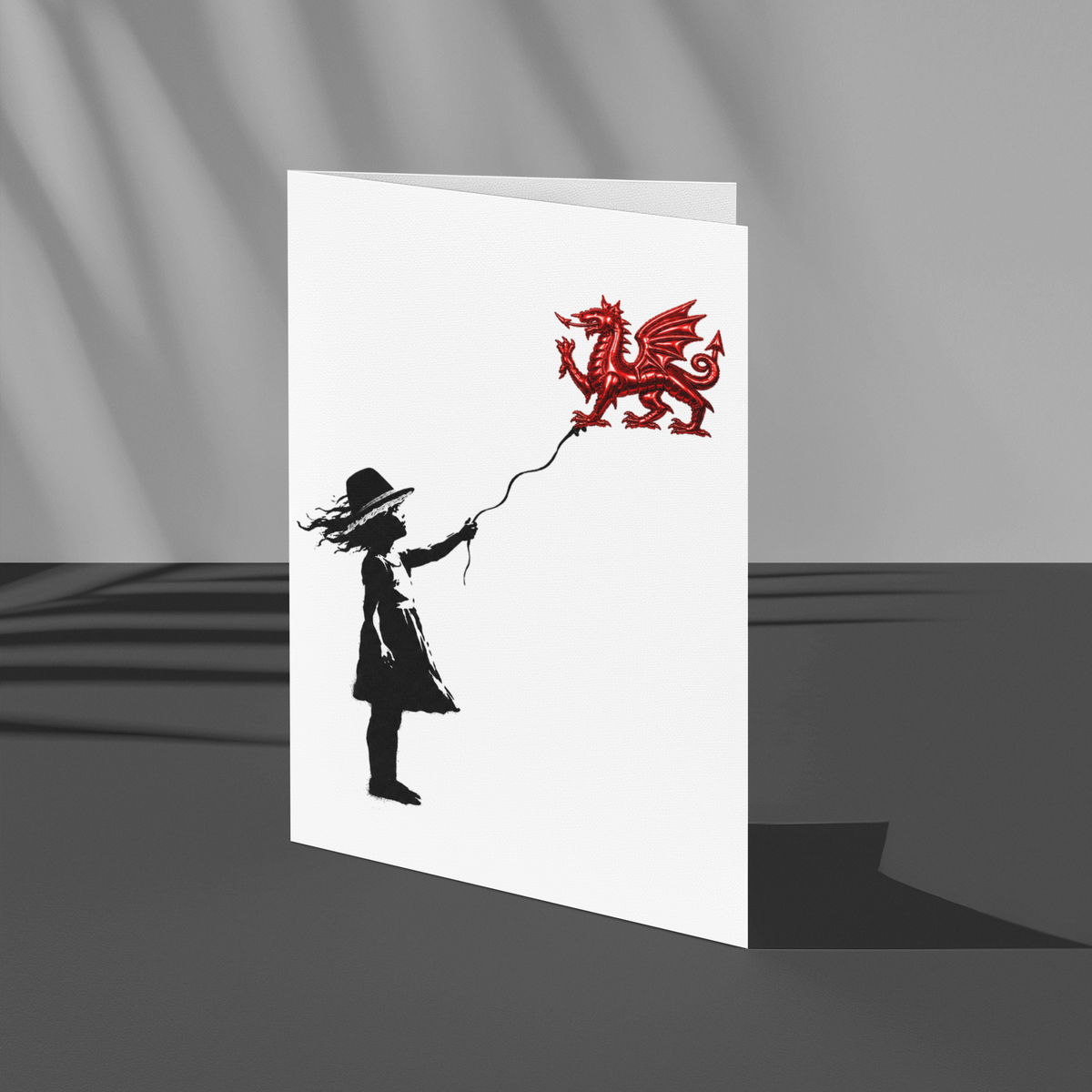 A white greeting card featuring a black silhouette of a child holding a red dragon balloon, styled standing upright on a dark gray surface with dramatic diagonal shadows.