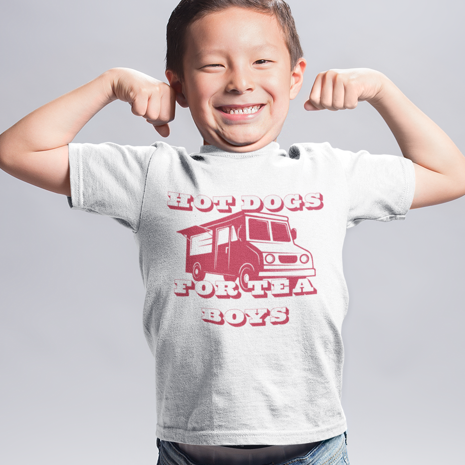 A young boy smiling and flexing his biceps, wearing a light gray graphic t-shirt with a red truck design and the text 'HOT DOGS FOR TEA BOYS', blue jeans, and blue sneakers, against a light gray studio background.
