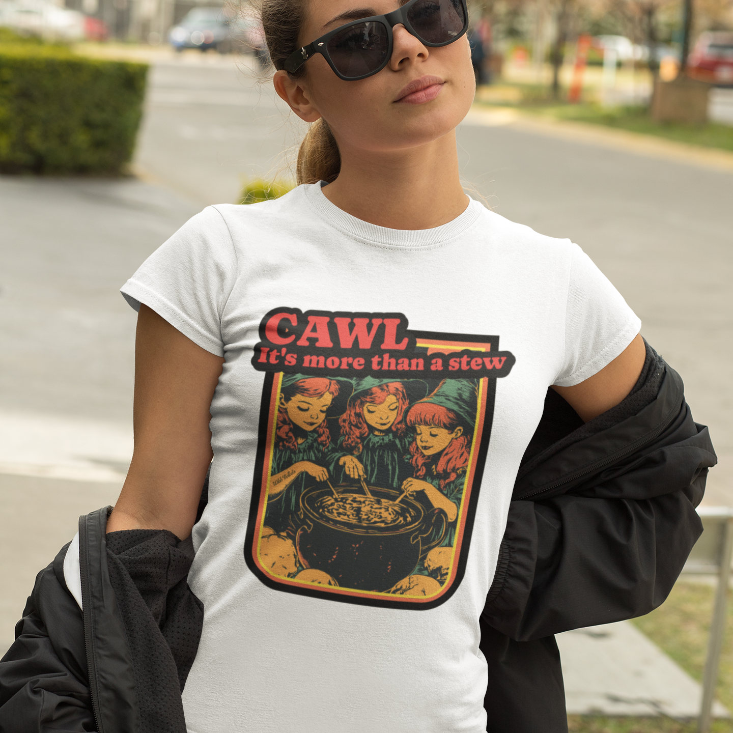 A woman wearing a white graphic t-shirt with a 'CAWL it's more than a stew' print, styled with a black jacket draped off her shoulders and sunglasses, set against an outdoor street background.