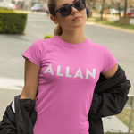 A woman wearing a bright pink crewneck t-shirt with the name 'ALLAN' printed in white across the chest, styled with a black jacket draped off her shoulders and black sunglasses, set against an outdoor street background.
