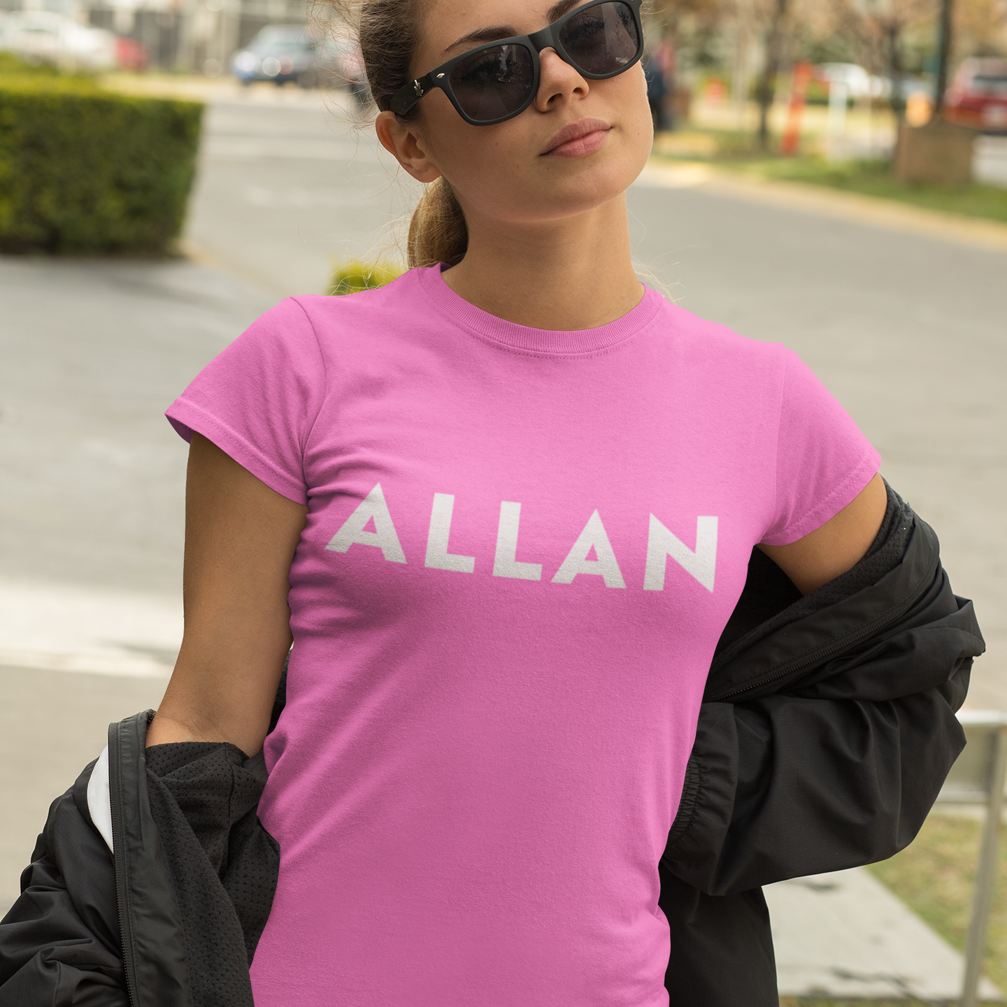 A woman wearing a bright pink crewneck t-shirt with the name 'ALLAN' printed in white across the chest, styled with a black jacket draped off her shoulders and black sunglasses, set against an outdoor street background.