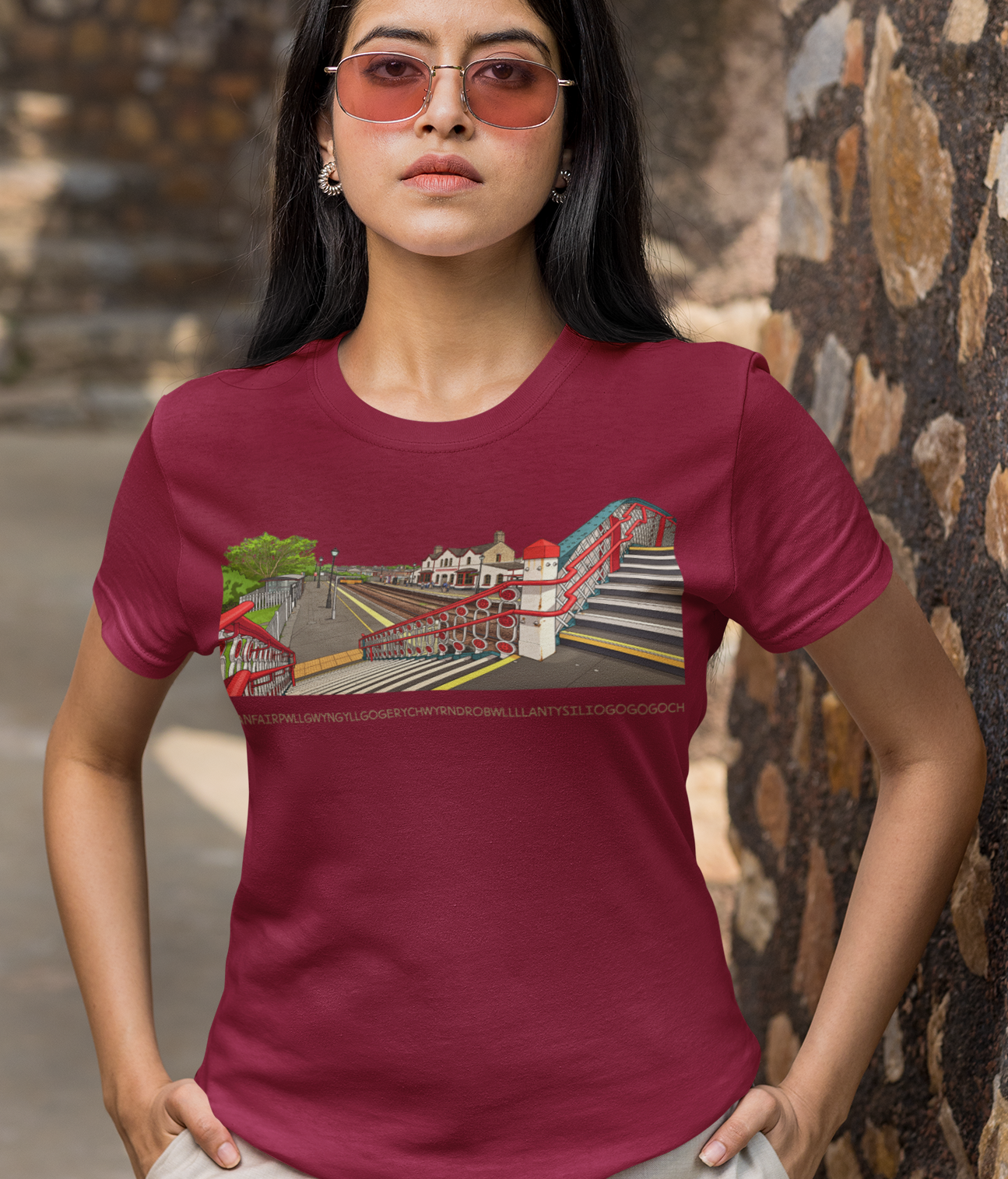 A woman wearing a maroon short-sleeve graphic t-shirt and beige pants, standing outdoors against a stone wall.