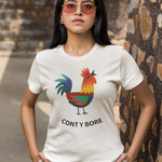 A woman wearing a cream-colored graphic t-shirt with a colorful rooster illustration and the text 'CONT Y BORE' printed below it. She is posing outdoors against a rustic stone wall.