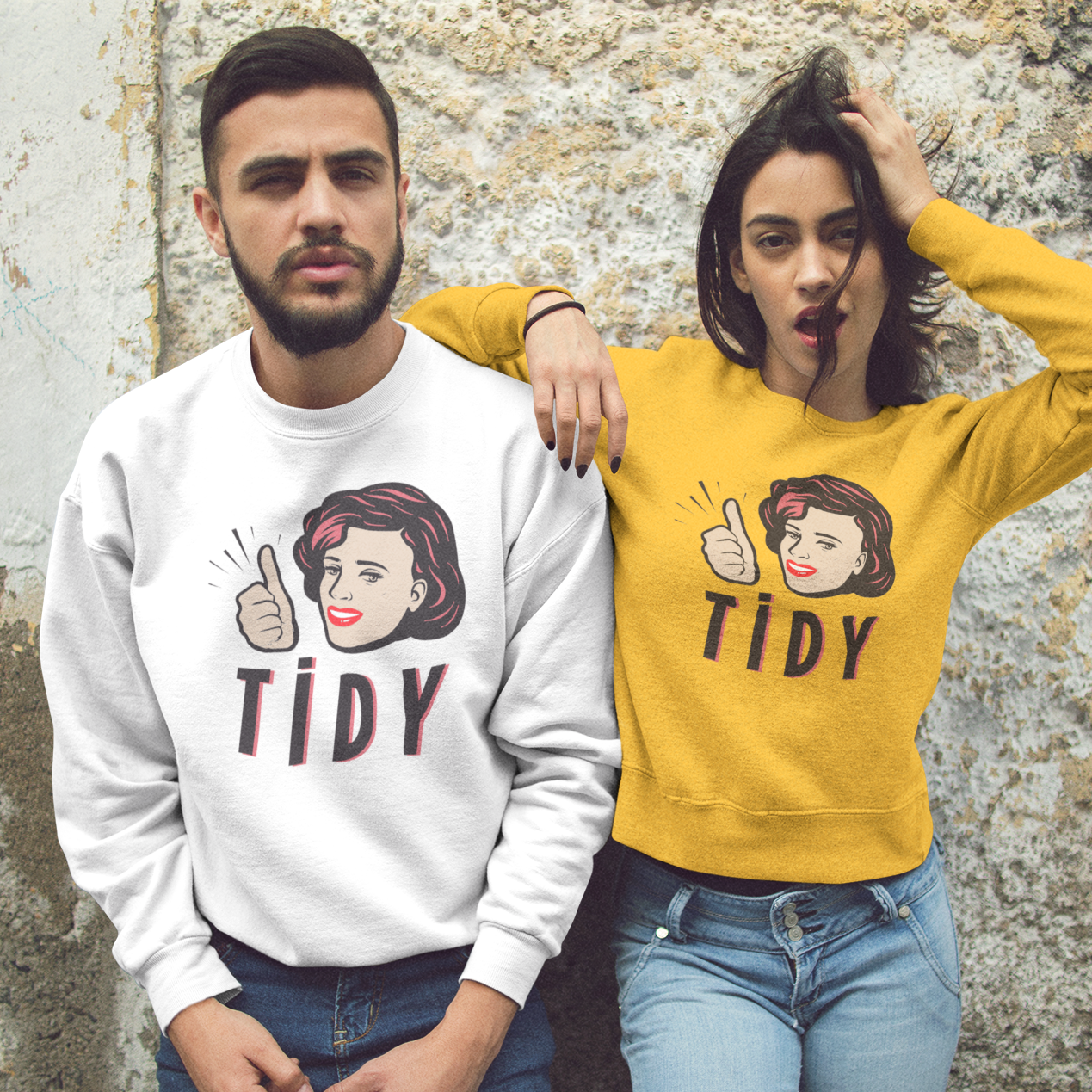 Two models posing against a textured stone wall, wearing matching crewneck sweatshirts. The model on the left wears a white sweatshirt with a cartoon graphic of a woman giving a thumbs-up and the word 'TIDY' in dark red.