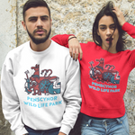 A man and a woman standing against a textured stone wall, wearing matching crewneck sweatshirts. The man wears a white sweatshirt with the Penscynor wild life park graphic, and the woman wears a red sweatshirt withthe same.