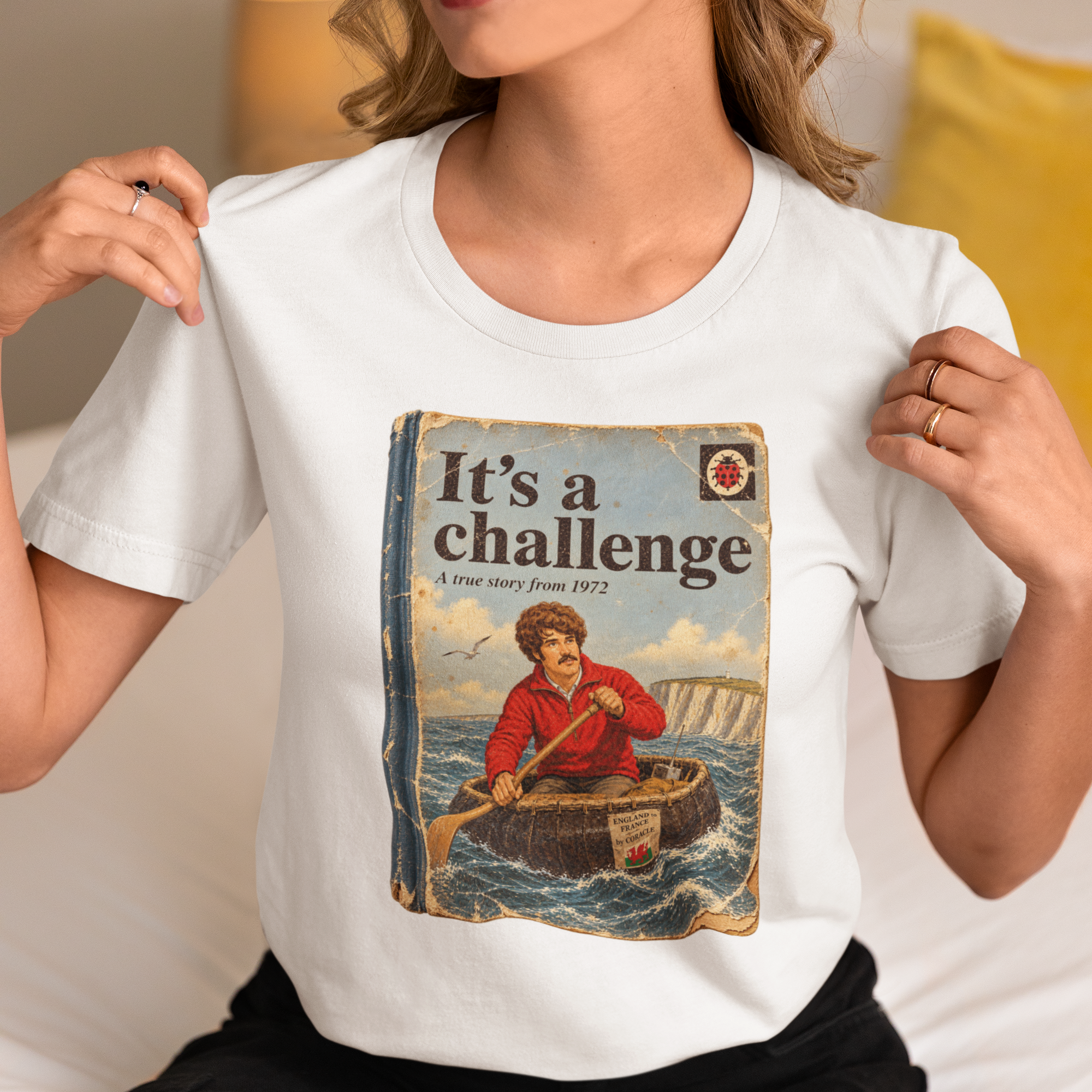 A woman wearing a white graphic t-shirt with a vintage-style book cover print that reads 'It's a challenge' and shows the product in a cozy indoor setting.
