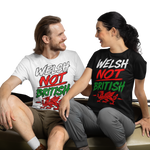 A man and a woman sit on a beige kayak placed on artificial grass, wearing matching graphic T-shirts that read 'WELSH NOT BRITISH' in white, red, and green. The background is a clean white studio backdrop.