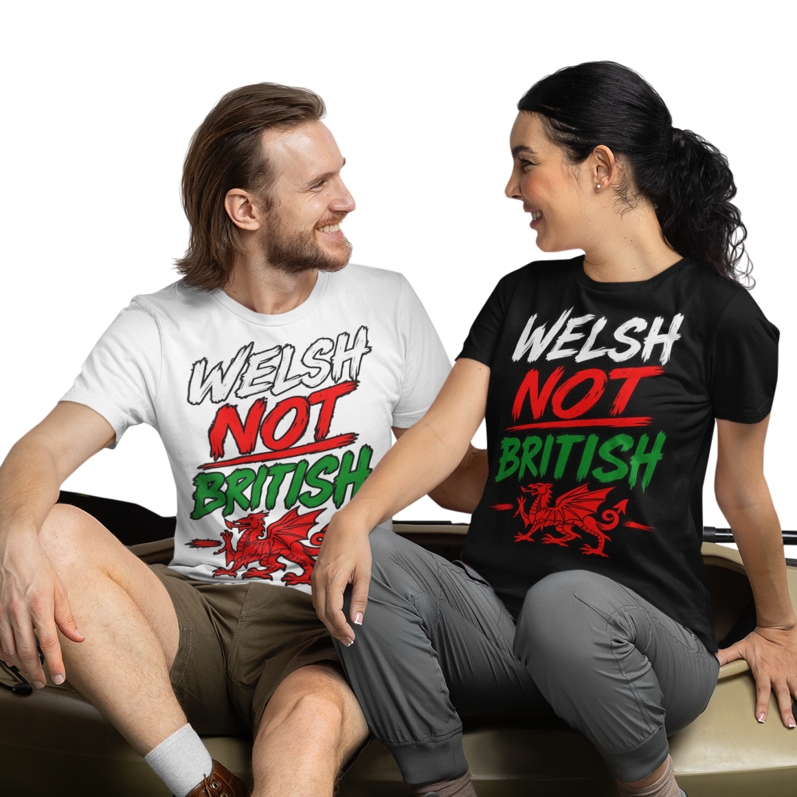 A man and a woman sit on a beige kayak placed on artificial grass, wearing matching graphic T-shirts that read 'WELSH NOT BRITISH' in white, red, and green. The background is a clean white studio backdrop.
