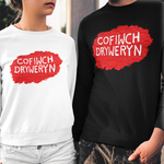 Two models wearing matching crewneck sweatshirts with a red paint-splatter graphic and the Welsh phrase 'COFIWCH DRYWERYN' in white text. The model on the left wears a white sweatshirt and denim shorts, while the model on the right wears a black sweatshirt and distressed black jeans.