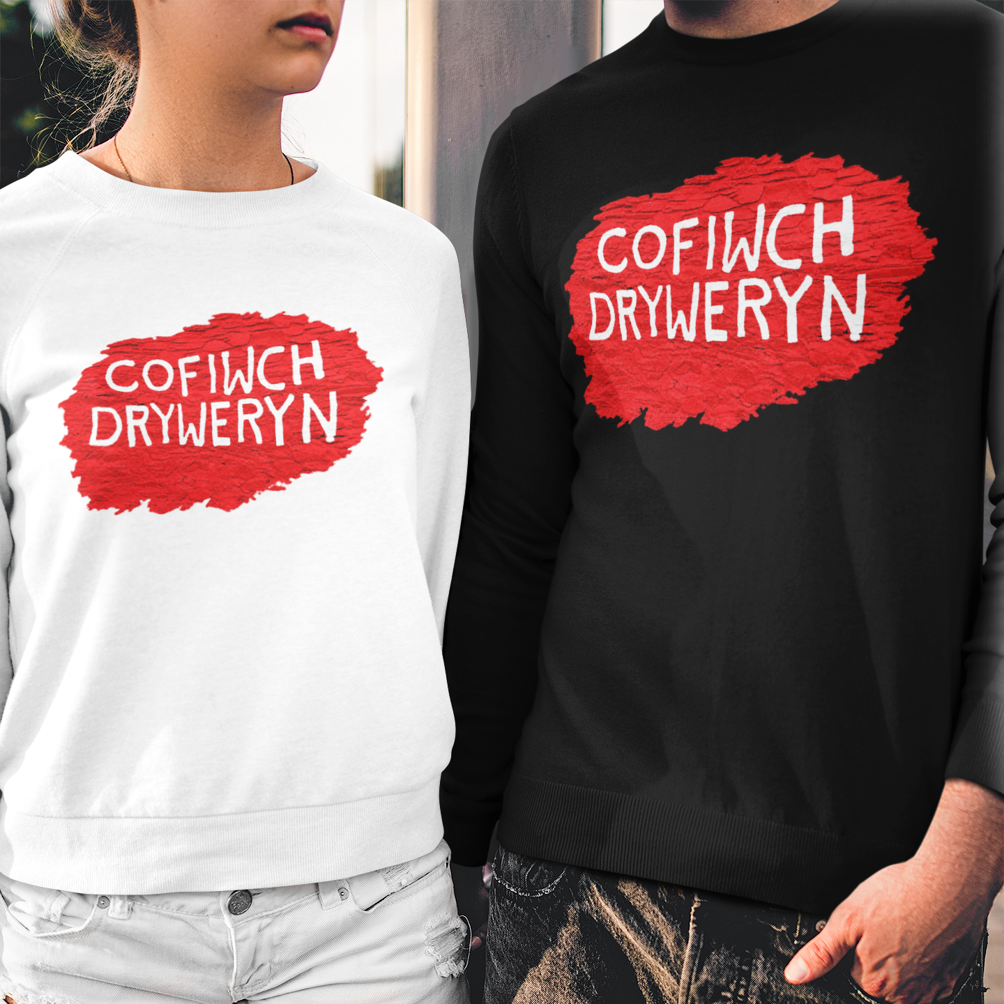 Two models wearing matching crewneck sweatshirts with a red paint-splatter graphic and the Welsh phrase 'COFIWCH DRYWERYN' in white text. The model on the left wears a white sweatshirt and denim shorts, while the model on the right wears a black sweatshirt and distressed black jeans.