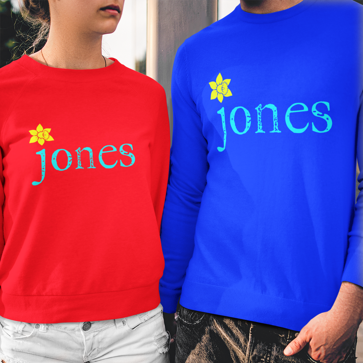 Two models wearing matching crewneck sweatshirts with 'jones' printed in teal with a yellow flower accent, one in red and one in blue, paired with white denim shorts and distressed black jeans respectively.