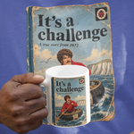 A person wearing a blue T-shirt holds up a white ceramic mug printed with a vintage-style book cover titled "It's a challenge" featuring a man in a boat.