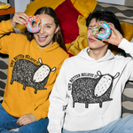 Two people sitting on a couch wearing matching graphic hoodies, each holding a donut over one eye. The person on the left wears a mustard yellow hoodie, and the person on the right wears a white hoodie.