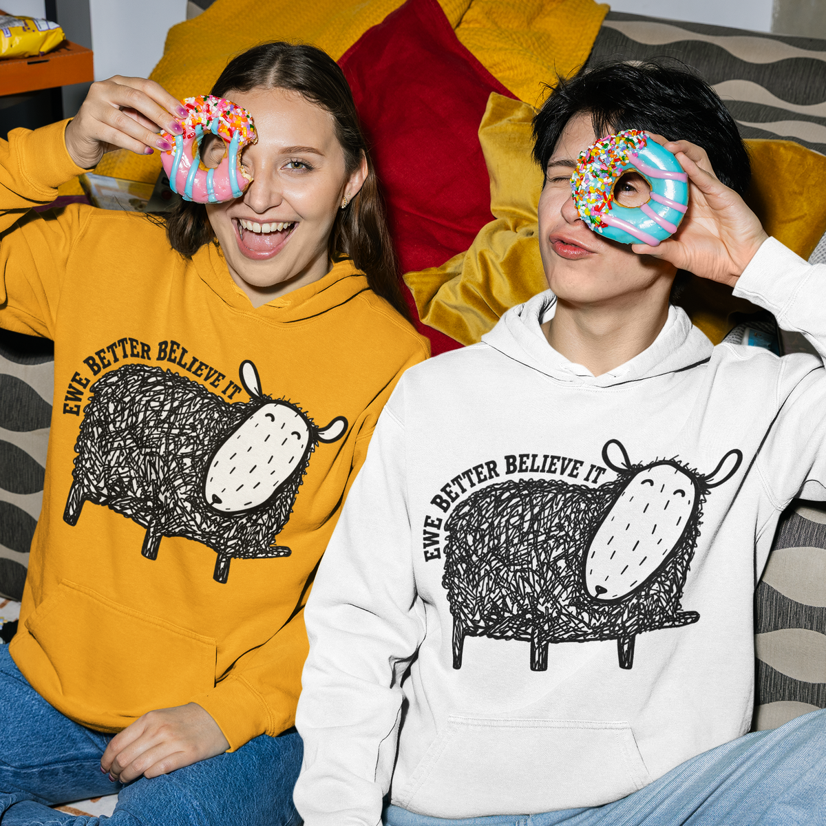 Two people sitting on a couch wearing matching graphic hoodies, each holding a donut over one eye. The person on the left wears a mustard yellow hoodie, and the person on the right wears a white hoodie.