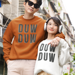 A couple posing on a city street wearing matching oversized sweatshirts with 'DUW DUW' printed on the front. The man wears an orange-brown sweatshirt and beige pants, while the woman wears a white sweatshirt, a beige pleated skirt, and white headphones.