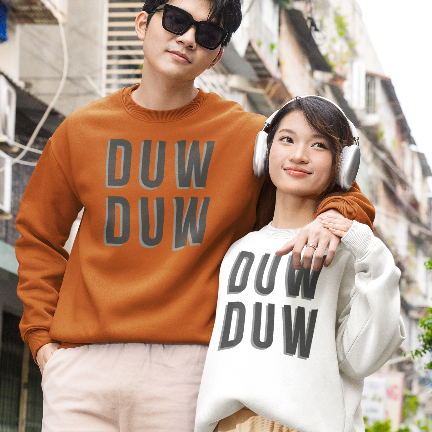 A couple posing on a city street wearing matching oversized sweatshirts with 'DUW DUW' printed on the front. The man wears an orange-brown sweatshirt and beige pants, while the woman wears a white sweatshirt, a beige pleated skirt, and white headphones.