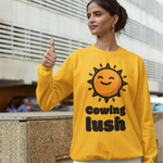 A woman wearing a bright yellow crewneck sweatshirt with a smiling sun graphic and the text 'Cowing lush' stands outdoors in front of a modern building, giving a thumbs-up.