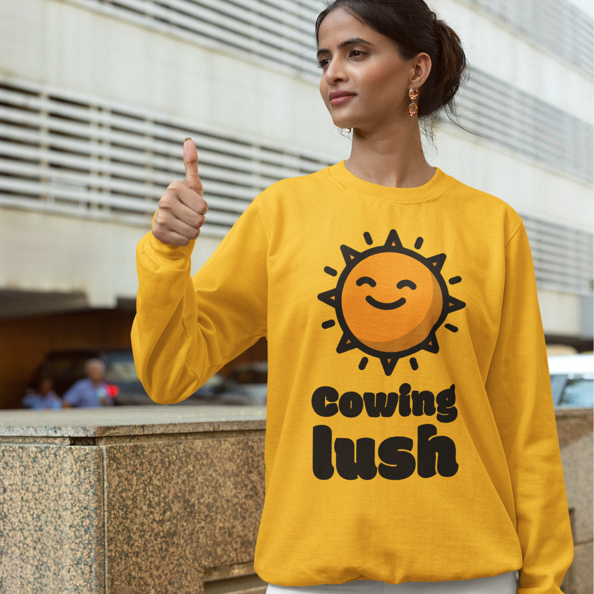 A woman wearing a bright yellow crewneck sweatshirt with a smiling sun graphic and the text 'Cowing lush' stands outdoors in front of a modern building, giving a thumbs-up.