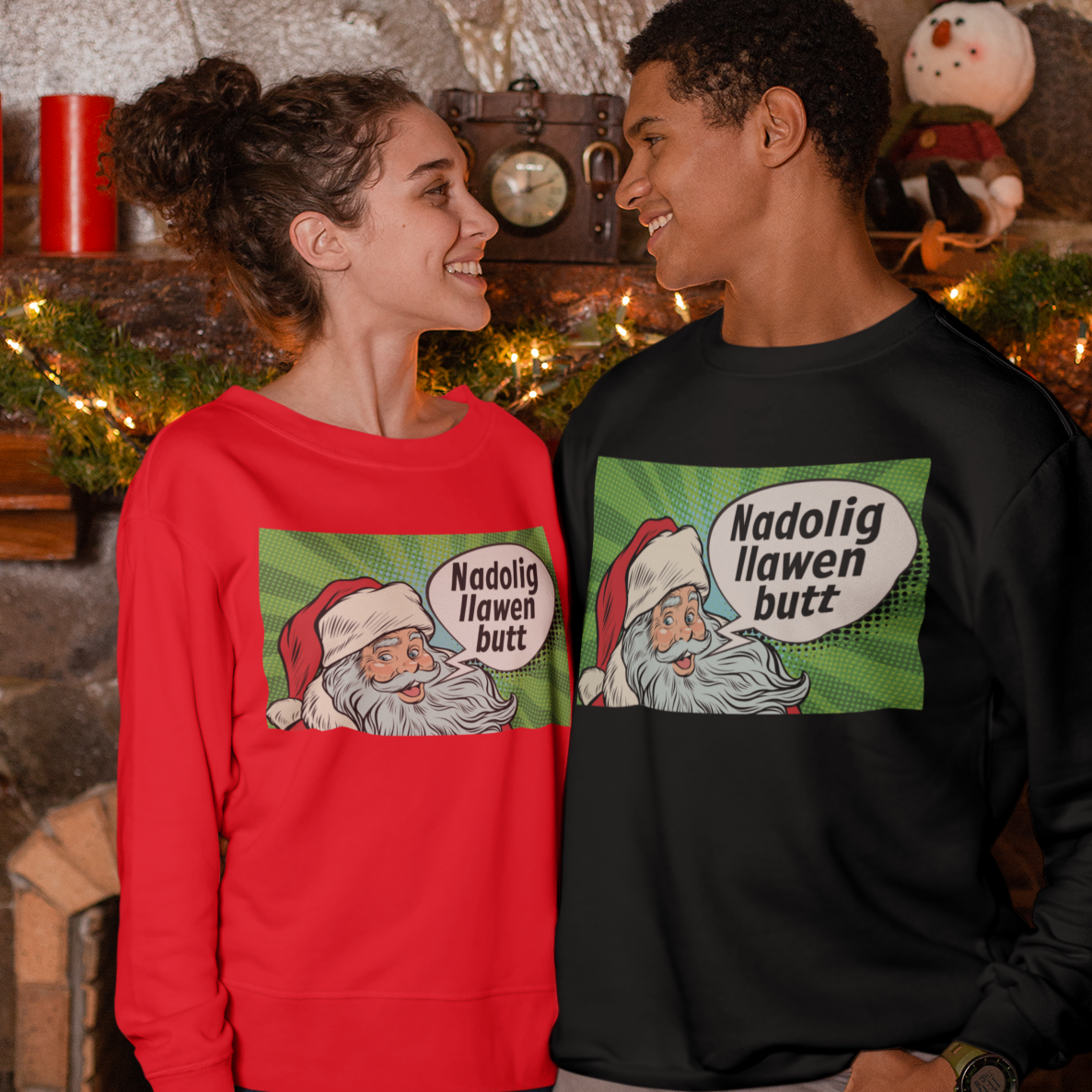 A man and a woman standing in front of a fireplace wearing matching Christmas sweatshirts with a cartoon Santa graphic and the text 'Nadolig Ilawen butt'.