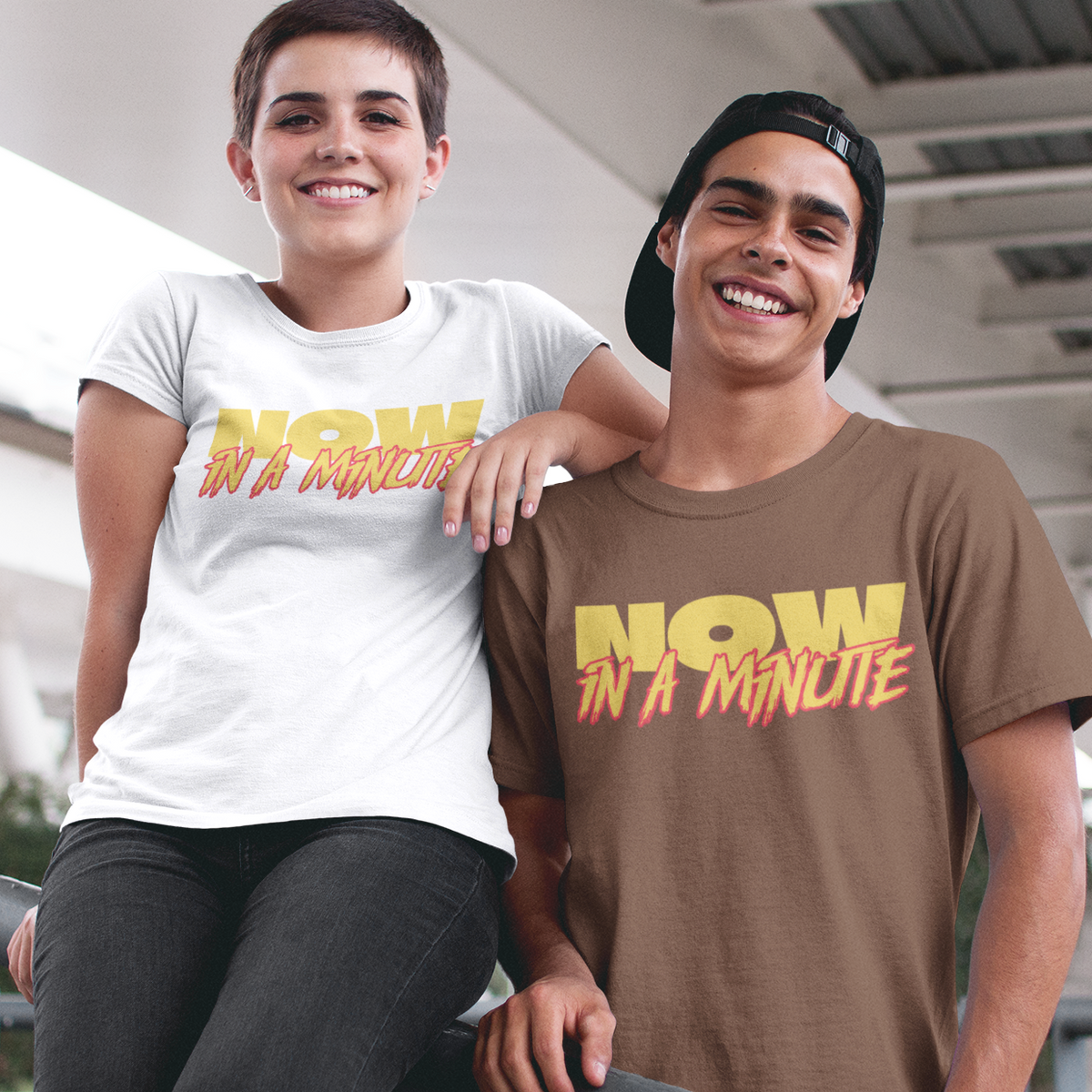 Two young men smiling and posing together on an outdoor bridge or overpass. The person on the left wears a white graphic T-shirt with yellow and orange text reading 'NOW IN A MINUTE', while the person on the right wears a brown graphic T-shirt with the same text in yellow and orange.