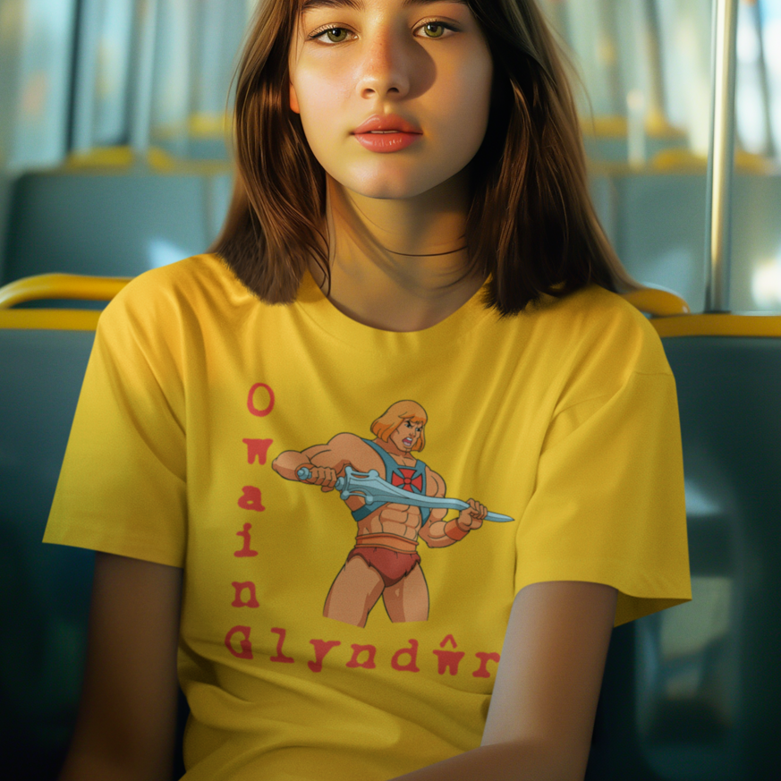 A young woman with brown hair sits on a bus wearing a bright yellow graphic t-shirt featuring He-Man and the text 'Owain Glyndŵr'.