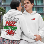 Two models sitting on a concrete bench outdoors, showcasing a white hoodie from both the front and back. The front view shows a small red graphic on the left chest, while the back view shows a large graphic reading 'WELSH NOT BRITISH' in white, red, and green.