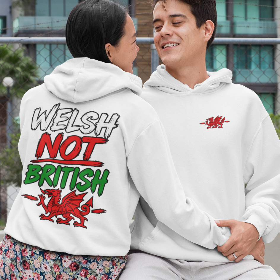 Two models sitting on a concrete bench outdoors, showcasing a white hoodie from both the front and back. The front view shows a small red graphic on the left chest, while the back view shows a large graphic reading 'WELSH NOT BRITISH' in white, red, and green.