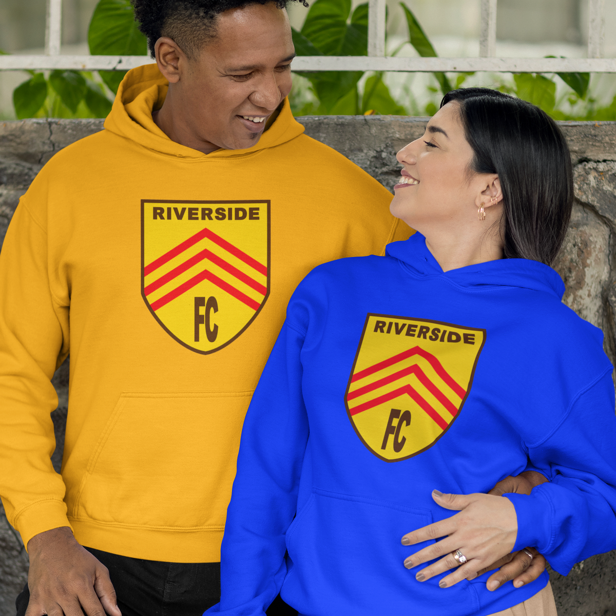 A man and a woman wearing matching hoodies with a 'RIVERSIDE FC' crest graphic. The man wears a golden-yellow hoodie, and the woman wears a royal blue hoodie.
