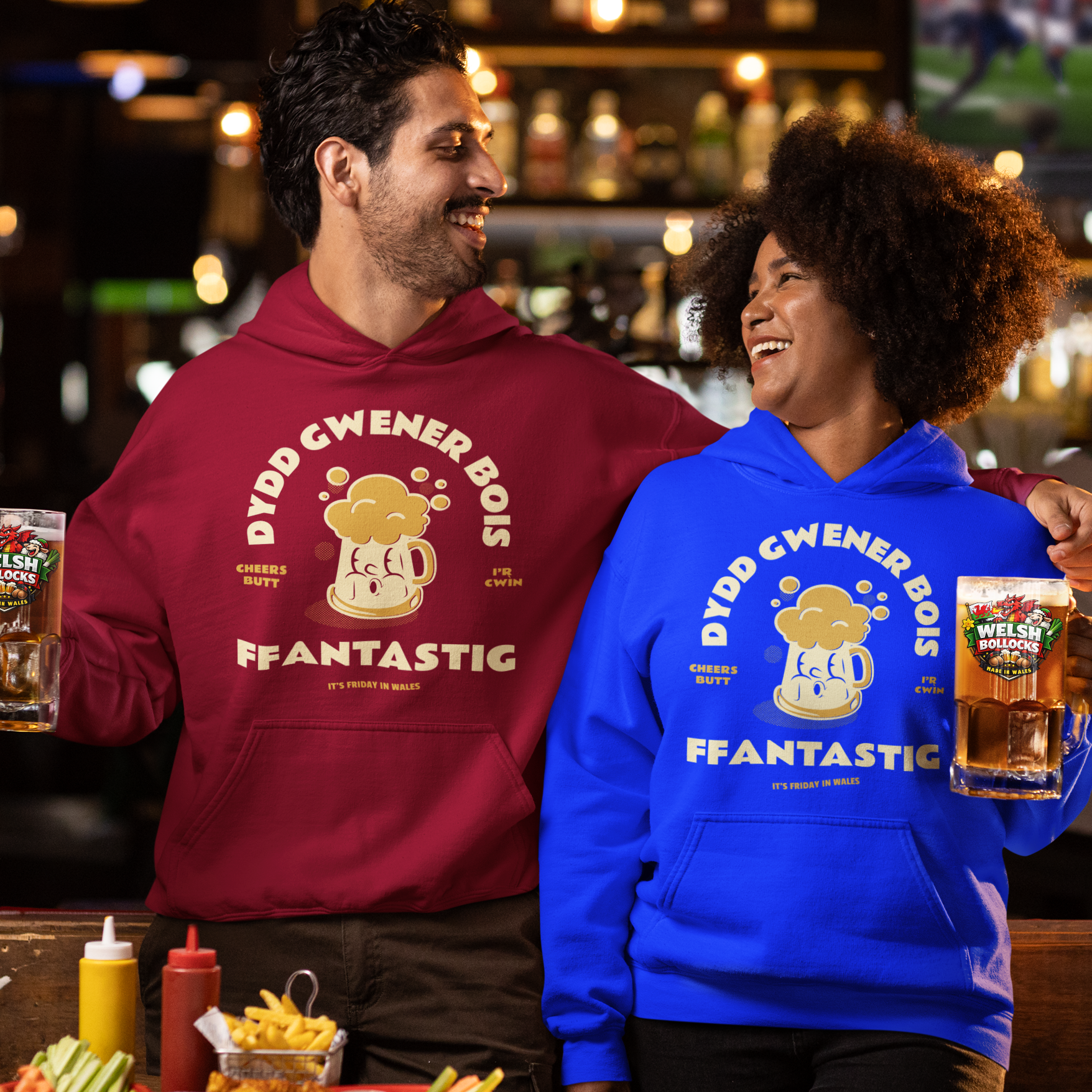 Two people standing in a bar setting, each wearing a colorful hoodie with a cartoon bear and the text 'DYDD GWENER BOIS FFANTASTIG' and holding a beer mug package. The person on the left wears a maroon hoodie, and the person on the right wears a bright blue hoodie.