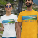 A man and a woman hiking hand-in-hand on a mountain trail, wearing matching graphic t-shirts. The man's shirt is bright yellow, and the woman's shirt is white, both featuring a scenic landscape print of a lake and beach with the text 'LLANDUNO'.