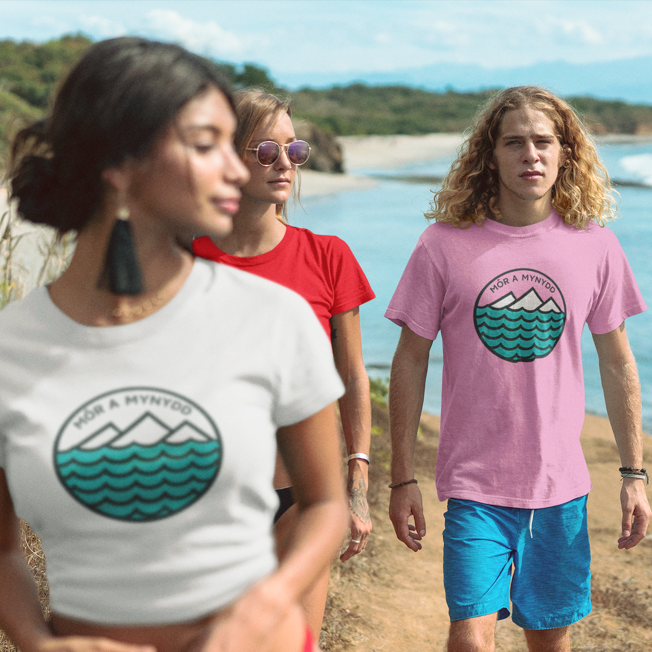 Three people wearing t-shirts with a circular logo featuring mountains and waves, standing on a beach.