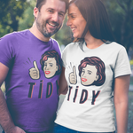 A man and a woman standing outdoors in a sunny park-like setting, both wearing matching graphic T-shirts. The man wears a purple T-shirt with a cartoon woman's face giving a thumbs-up and the word 'TIDY' in dark red letters.