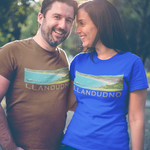 A man and a woman standing outdoors in a sunny park-like setting, both wearing graphic t-shirts. The man wears a brown t-shirt and the woman wears a bright blue t-shirt, both featuring a landscape graphic and the text 'LLANDUDNO'.