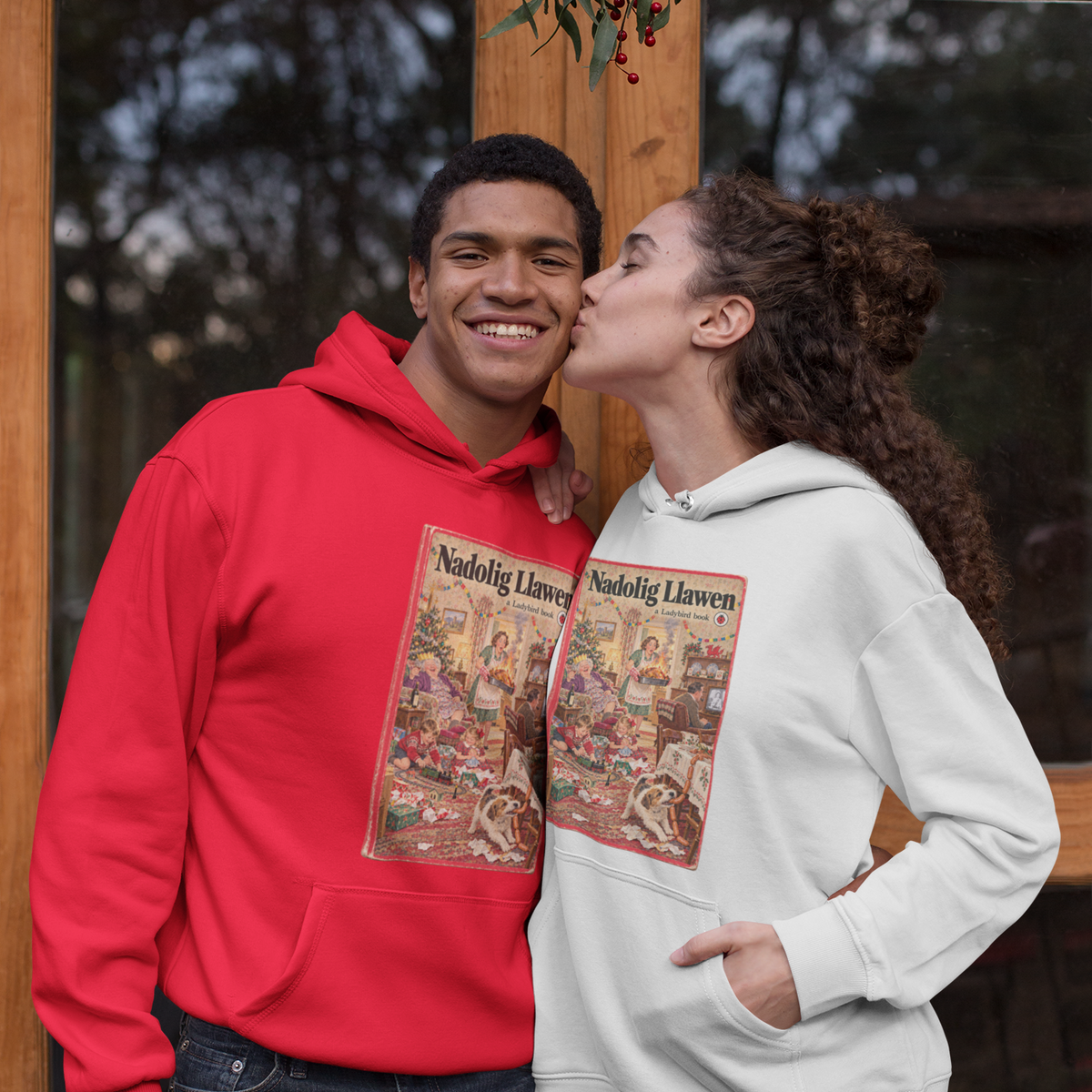 Two young adults posing in front of a rustic wooden door. The person on the left wears a bright red hoodie with a vintage Christmas card graphic on the front, and the person on the right wears a light gray hoodie with a matching graphic.