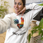 A woman takes a mirror selfie wearing a white hoodie with a colorful rooster graphic and the text 'CONT Y BORE' printed on the front, paired with blue jeans in a home setting with plants.