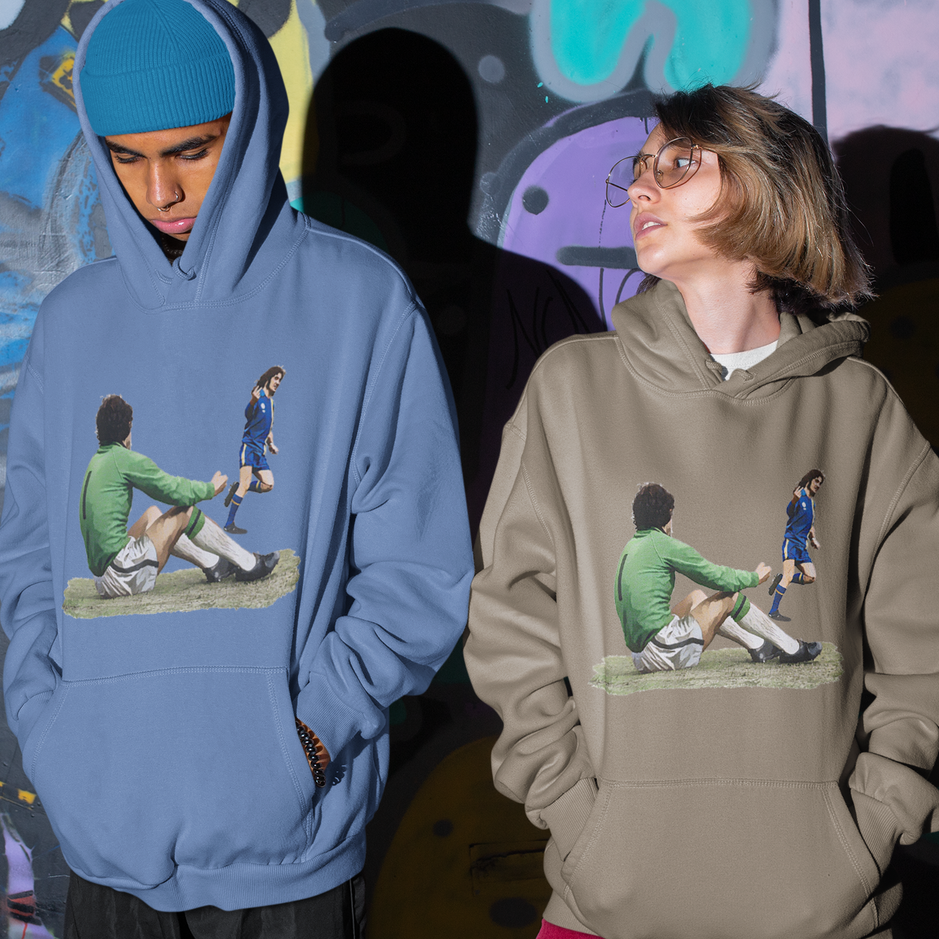 Two people wearing hoodies with graphic designs in front of a graffiti wall.