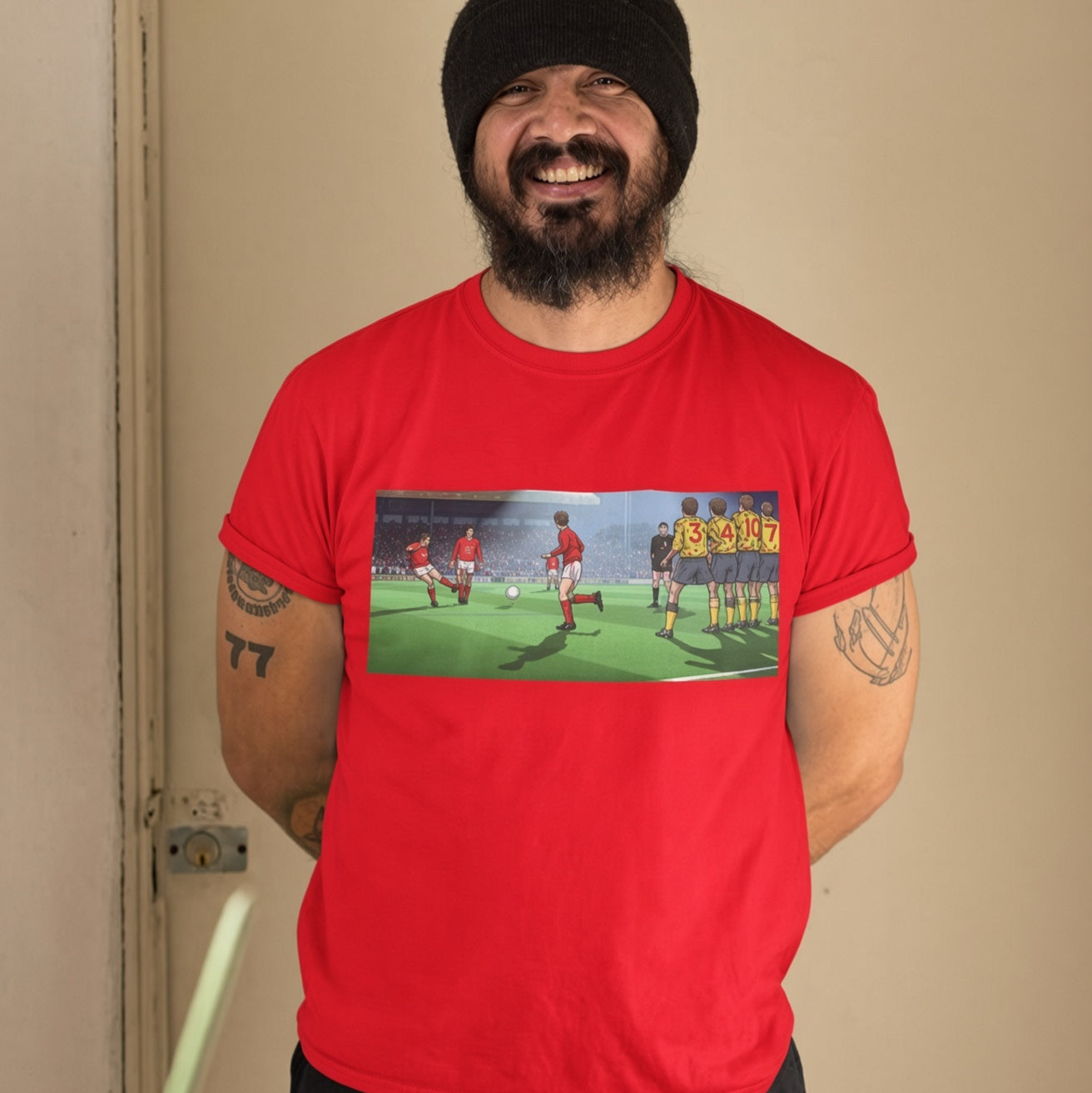 A smiling man wearing a black beanie and a bright red graphic t-shirt. The t-shirt features a wide rectangular print on the chest depicting a soccer scene with players and a scoreboard showing Mickey Thomas scoring the equaliser against Arsenal in January 1992.