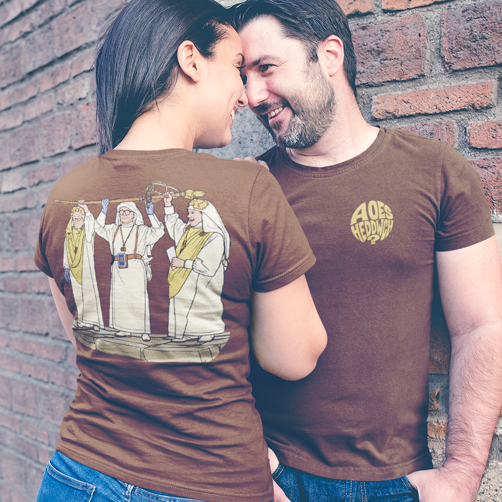 A couple wearing matching brown graphic t-shirts posing against a red brick wall. The woman shows the back of her shirt with a detailed illustration, while the man shows a gold circular logo on the chest.
