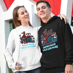 A man and a woman standing outdoors in front of a modern red and white geometric building, wearing matching hoodies. The woman's hoodie is white with a colorful graphic and the text 'PENSCTNOR WILD LIFE PARK', and the man's hoodie is black with the same graphic and text.