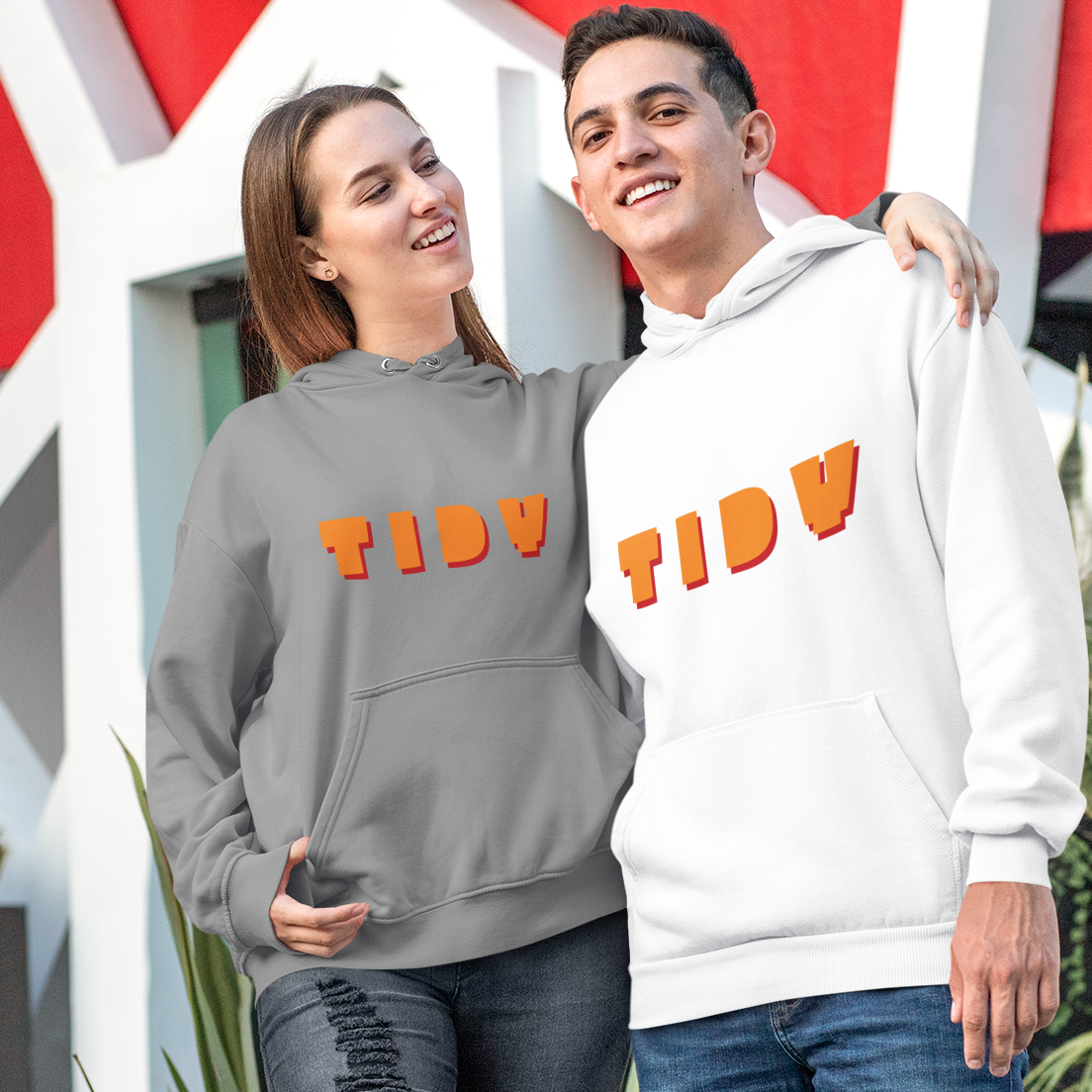 A man and a woman standing outdoors in front of a modern white and red geometric building. The woman wears a gray hoodie with orange 'TIDY' lettering, and the man wears a white hoodie with orange 'TIDY' lettering.