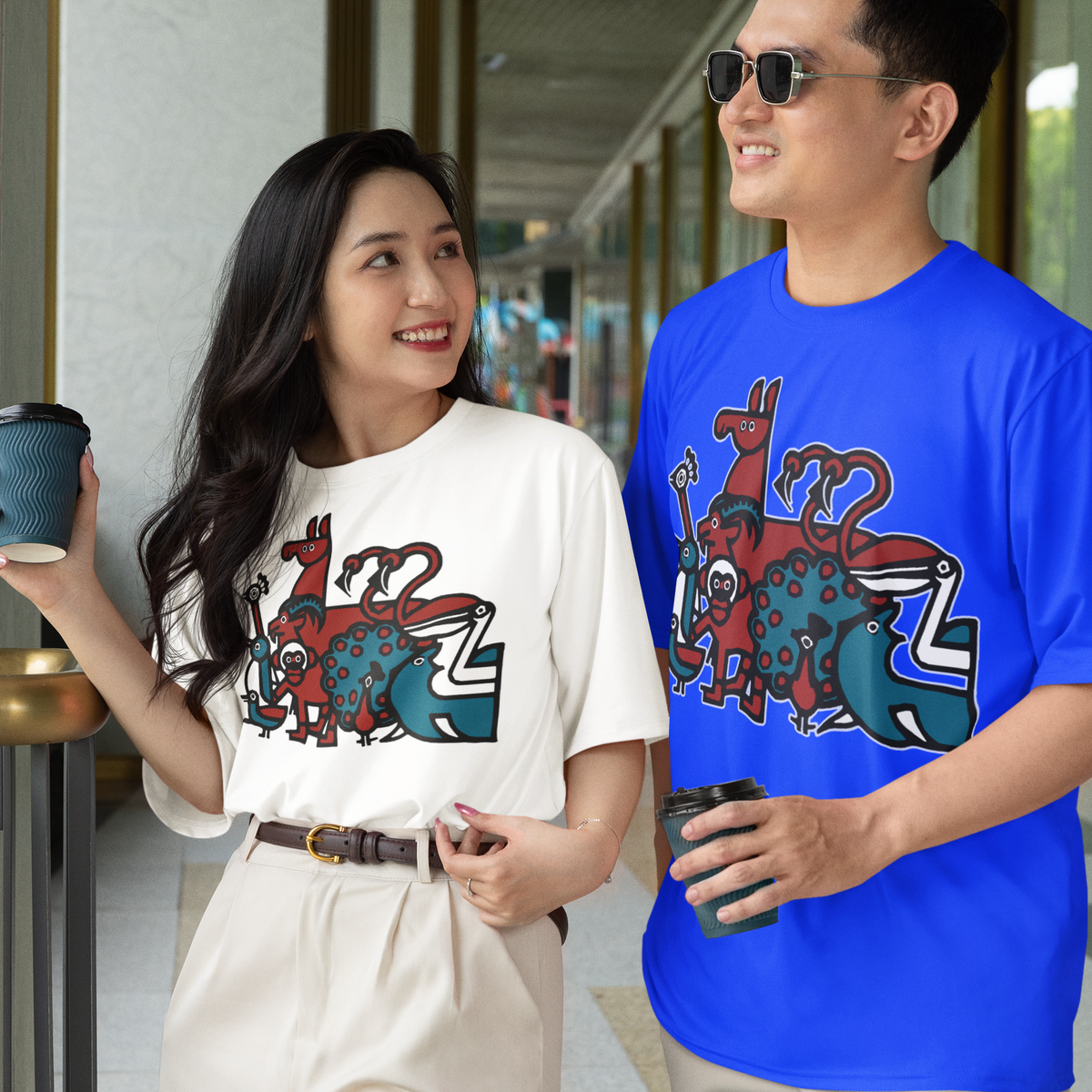 A man and a woman walking side-by-side in a modern corridor, both wearing matching graphic t-shirts. The woman's shirt is off-white with a colorful graphic of the Penscynor logo and cartoon elements, while the man's shirt is a vibrant royal blue with the same graphic.