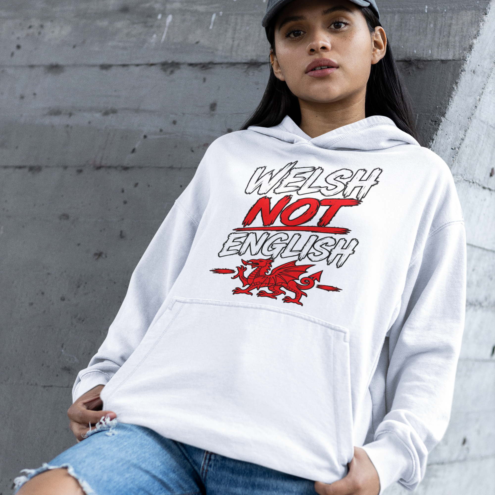 A woman wearing a white pullover hoodie with a 'Welsh Not English' graphic in red and black, paired with ripped blue jeans and a dark gray baseball cap, leaning against a gray concrete wall.
