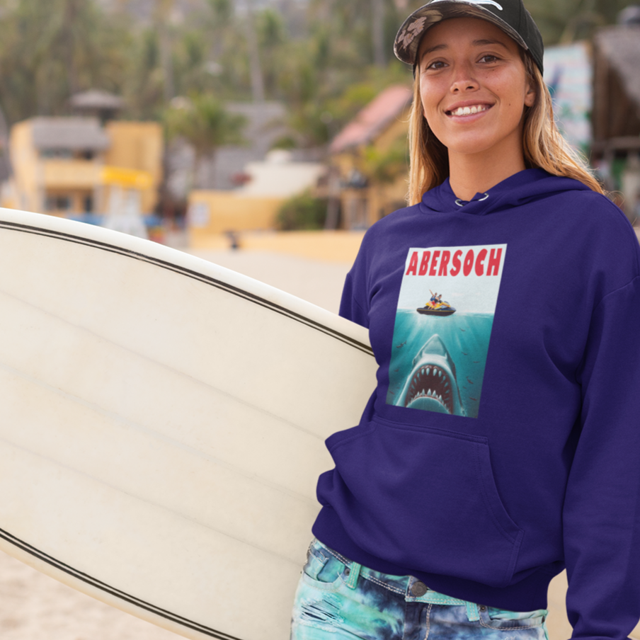 Woman holding a surfboard on a beach wearing a purple hoodie with a shark graphic.