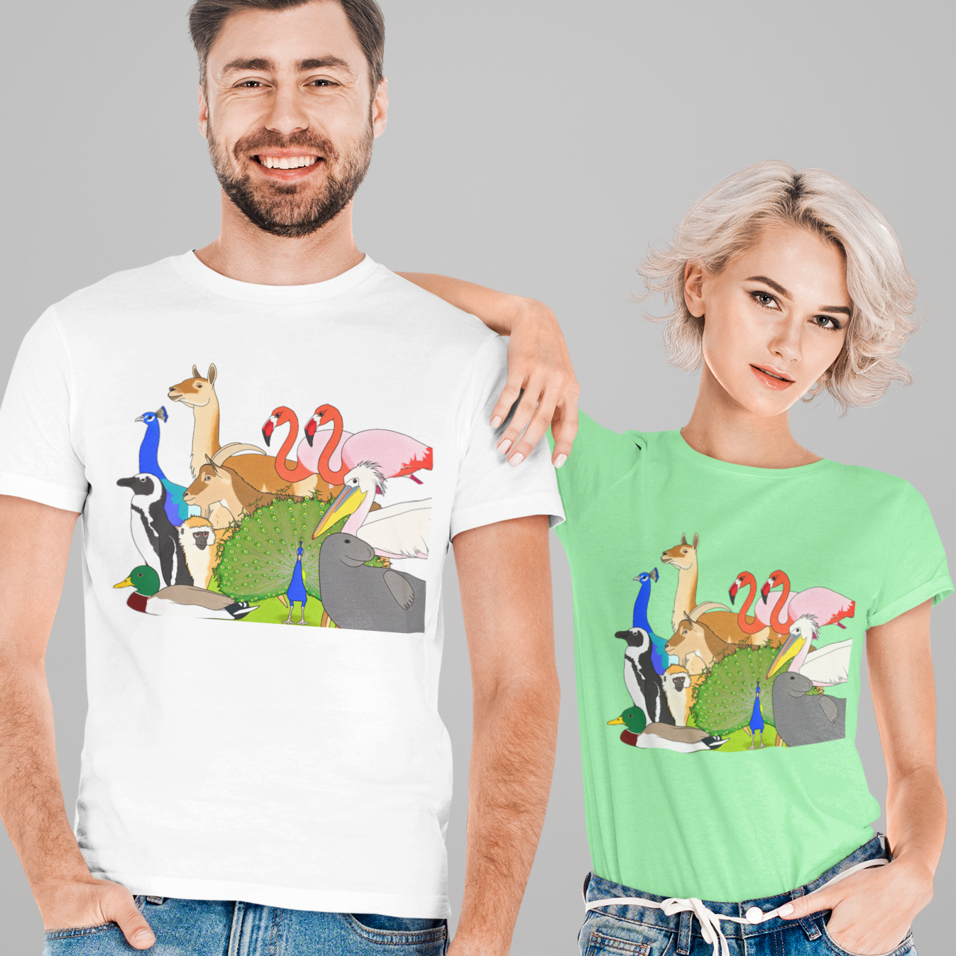 A man and a woman modeling matching graphic t-shirts against a solid gray background. The man wears a white t-shirt and the woman wears a mint green t-shirt, both featuring a colorful cartoon graphic of a giraffe, flamingos, and a green dinosaur.