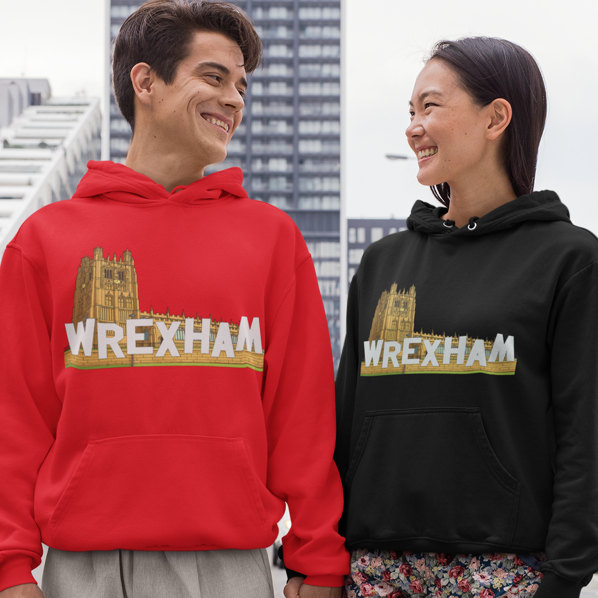 A man and a woman standing outdoors in a city setting, each wearing a pullover hoodie with a Wrexham graphic. The man's hoodie is bright red, and the woman's hoodie is black.