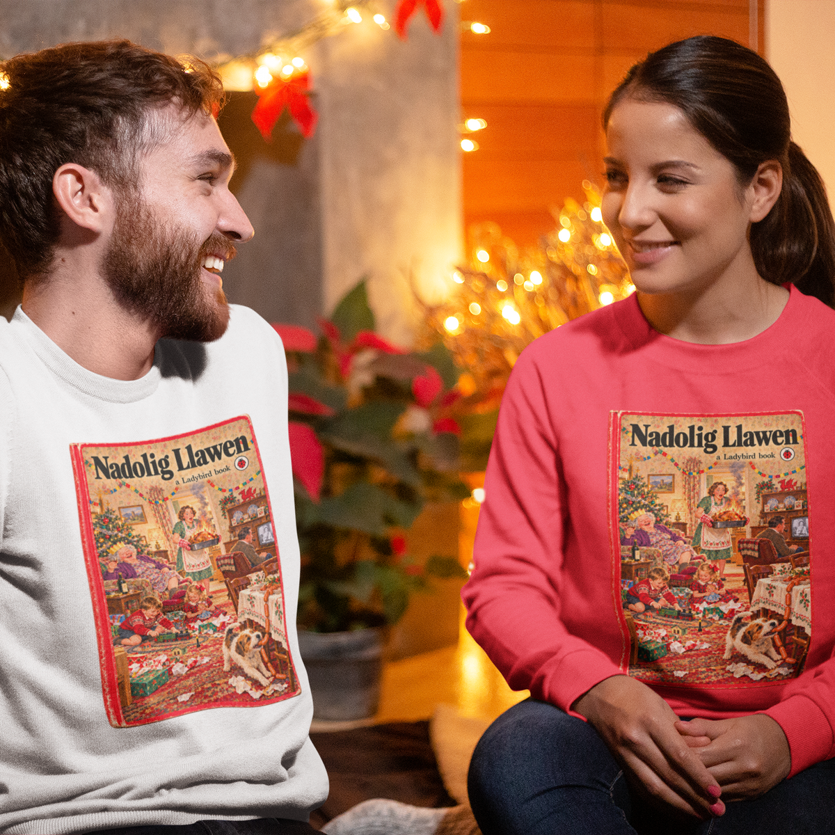 A man and a woman sitting side-by-side wearing matching crewneck sweatshirts with a colorful Christmas illustration of 'Nadolig Llawen' on the front. The man wears a light gray sweatshirt, and the woman wears a coral pink sweatshirt.