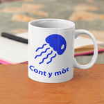 A white ceramic mug with a blue jellyfish graphic and the text 'Cont y mór' printed on it, sitting on a wooden table with a blurred bedroom background.